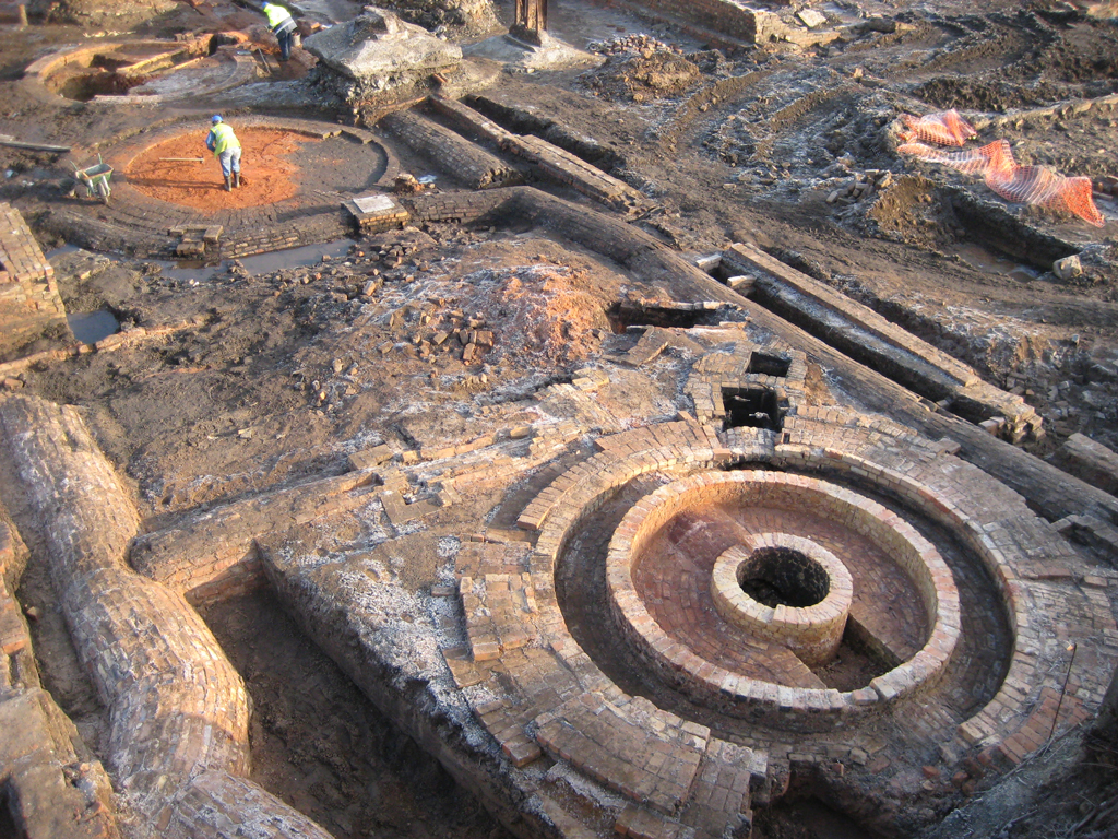 Archaeological dig with exposed circular brick structures, the kiln bases. A man is measurign one of them