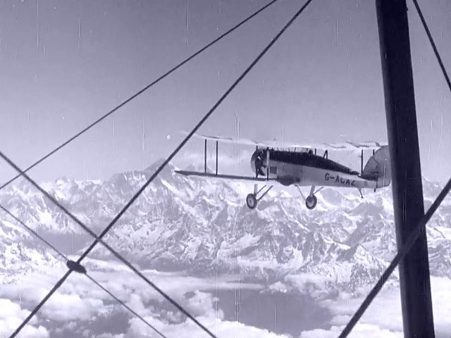 Period photo of biplane approaching snow covered mountain peaks, taken through the struts of the second plane