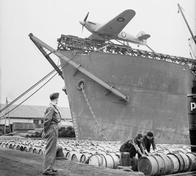 An airman watches as Algerian dock workers roll barrels of oil along the quayside at Algiers. Behind them is Hawker Sea Hurricane Mark I, W9182, of the Merchant Ship Fighter Unit mounted on the fo'c'sle catapult of a Catapult Armed Merchantman (CAM ship).