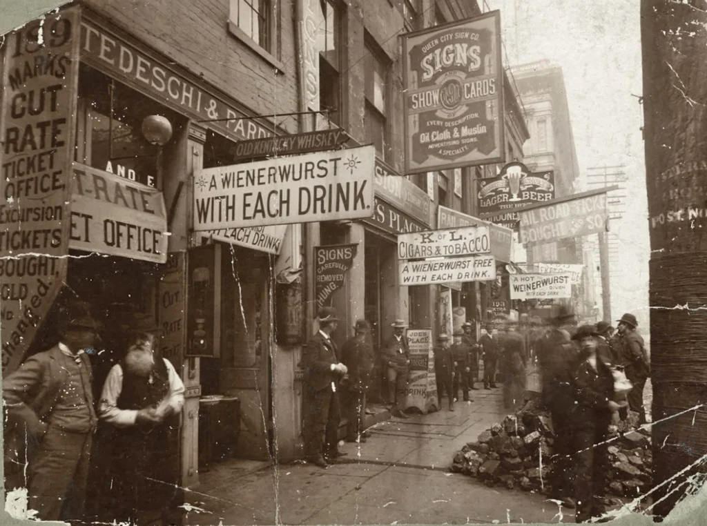 Old photo of a busy narrow street. Multiple shop signs read "A wienerwurst with every drink"