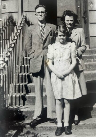 Steps of 20 Queen Square, with iron railings, much as today. Richard and Rosalind are adults, both in suits; Carol a teenager in a floral dress, stands in front of them