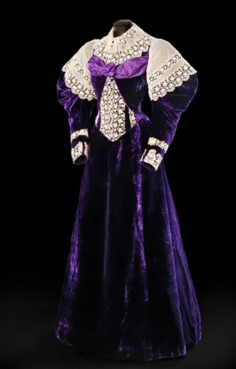 Woman’s dress, consisting of bodice (E.1973.57.e.1) and matching skirt (E.1973.57.e.2), in purple silk velvet with purple corded silk and cream machine-embroidered chemical lace with net bands. Bodice with small standing collar overlaid with deep falling frill of cream net and lace, fitted bodice with darts decorated with loosely arranged horizontal panel of purple corded silk across upper bust with ruched rosette at centre front, inverted v-shape at front covered in cream lace except for one stripe of velvet either side, fastening on left with eleven metal hooks and thread eyes to waist and five metal hooks and silk in flared basque, v-shaped waistline. Full-length gigot or leg-of-mutton sleeves with fullness pleated into shoulder overlaid with deep frill of cream lace, tapering into fitted cuff trimmed with narrow band of cream lace around lower arm and wider open-ended band at cuff. Back fitted with slight v-shaped waistline and small basque with curved hem.